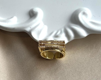 May include: A gold-colored ring with three rows of small, clear gemstones. The ring has an open design and is set against a white, decorative plate and a beige fabric background. The ring is a piece of jewelry.