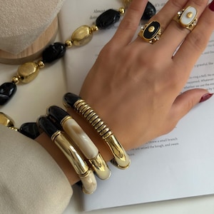 May include: Gold-toned jewellery set featuring rings, bracelets, and a necklace. The rings have black and white oval stones. Bracelets have black and white details. The necklace has black and gold beads.