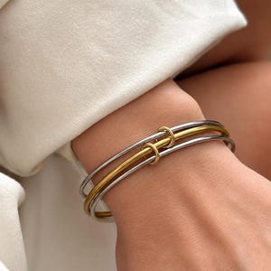 Peut inclure: Un bracelet jonc multi-rangs composé de deux anneaux fins en métal argenté et d'un anneau central en métal doré, le tout maintenu par deux petits anneaux circulaires dorés qui relient les trois bandes en un alignement fixe.