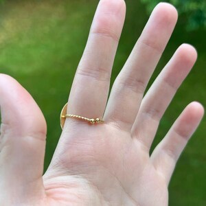 Puede incluir: Un anillo dorado con un peque&ntilde;o dise&ntilde;o plano y redondo. La banda del anillo tiene un dise&ntilde;o de cuerda trenzada con dos peque&ntilde;as cuentas doradas. El anillo est&aacute; en una mano con un fondo verde.