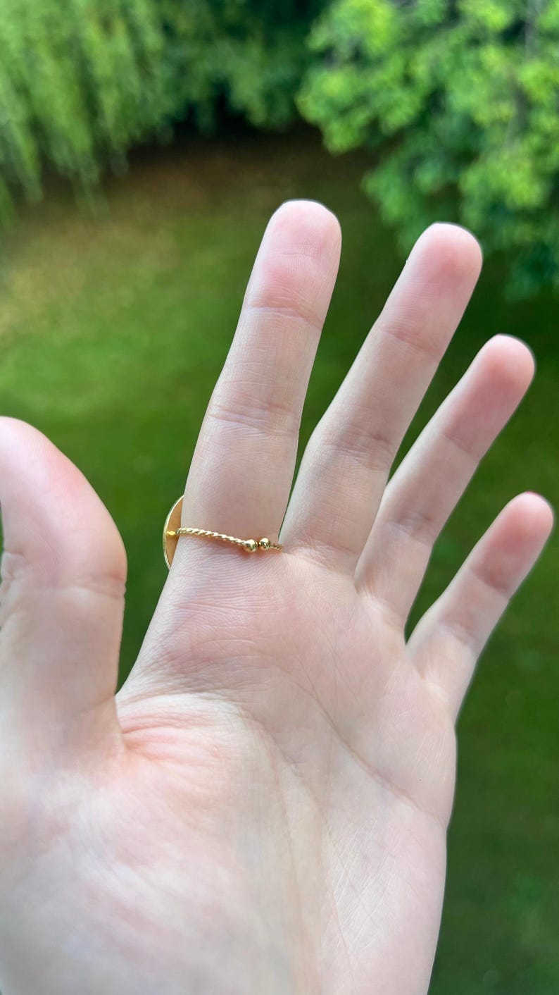 Puede incluir: Un anillo dorado con un peque&ntilde;o dise&ntilde;o plano y redondo. La banda del anillo tiene un dise&ntilde;o de cuerda trenzada con dos peque&ntilde;as cuentas doradas. El anillo est&aacute; en una mano con un fondo verde.