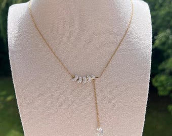 May include: A gold-toned necklace with a delicate design. The necklace features a series of small, sparkling embellishments in the center, with a single crystal pendant hanging below. The necklace is displayed on a mannequin.