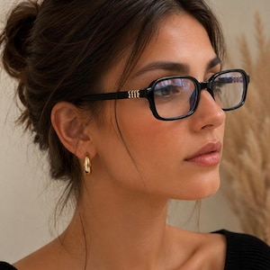 May include: Black rectangular spectacles featuring a glossy finish and clear lenses. The temples are accented with a small, metallic gold-toned decorative element near the hinge, providing a subtle contrast to the dark frame structure.