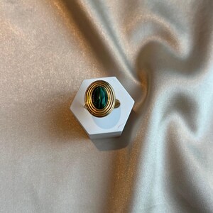 Puede incluir: Un anillo dorado con una piedra ovalada de color verde oscuro. El anillo presenta un dise&ntilde;o en capas alrededor de la piedra, colocado sobre un expositor hexagonal blanco. El fondo es una tela clara y texturizada.