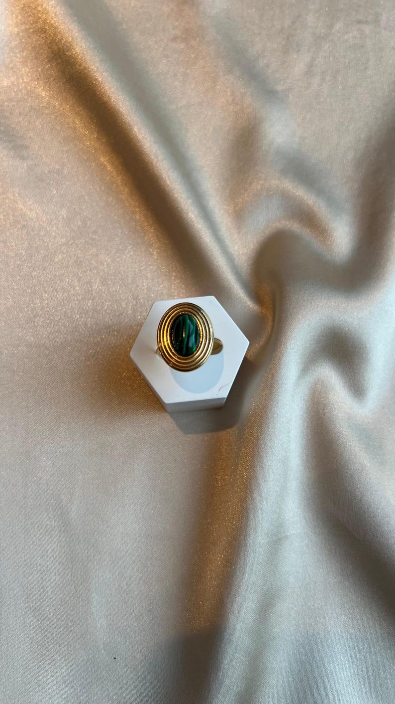 Puede incluir: Un anillo dorado con una piedra ovalada de color verde oscuro. El anillo presenta un dise&ntilde;o en capas alrededor de la piedra, colocado sobre un expositor hexagonal blanco. El fondo es una tela clara y texturizada.