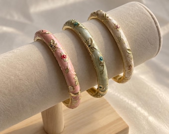 Könnte beinhalten: Drei aufklappbare Armreifen in Pastellrosa, Salbeigrün und Creme. Jeder goldfarbene Metallarmreif hat eine Emaille-Oberfläche mit geprägten goldenen Blumen- und Schmetterlingsmotiven, akzentuiert durch kleine bunte Steine.
