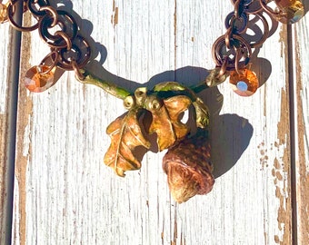 May include: A copper-toned necklace with a fall theme. The pendant features an acorn and leaves in shades of brown and gold. The necklace has copper chain links and amber-colored crystal accents.