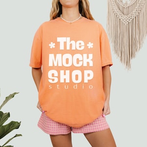 May include: A peach colored t-shirt with the text "*The* MOCK SHOP studio" printed in white. The model is wearing the t-shirt with pink and white checkered shorts.