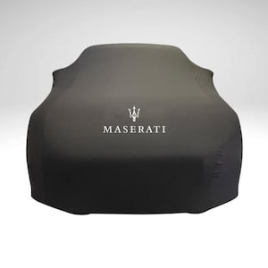 Puede incluir: Una funda de coche gris oscuro con el logotipo y el nombre de la marca Maserati en blanco. La funda está estirada sobre un coche, oscureciendo su forma. El fondo es un degradado de blanco a gris claro.