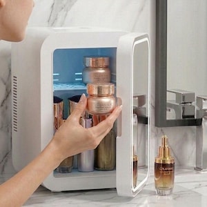 Könnte beinhalten: Ein weißer kompakter Hautpflege-Kühlschrank mit Innenbeleuchtung und Spiegeltür. Der Kühlschrank enthält mehrere Tiegel und Flaschen mit Hautpflegeprodukten, darunter ein roségoldener Tiegel mit der Aufschrift Cellavn Nu, sowie eine Flasche.