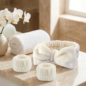 Op de afbeelding: Een set off-white pluche accessoires bestaande uit een brede hoofdband met een grote strik in het midden en twee bijpassende polsbandjes. De items hebben een zachte, pluizige textuur en een elastische constructie voor dagelijks gebruik.