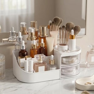 Può includere: Un organizer per cosmetici bianco in plastica con scomparti multipli e due cassetti trasparenti. L'organizer contiene vari flaconi per la cura della pelle, pennelli per il trucco, flaconi di profumo e piccoli contenitori per l'ordine.
