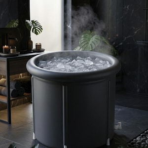 Peut inclure: Une baignoire cylindrique portable de couleur gris foncé pour bain froid, dotée d'un rebord épais et arrondi ainsi que de pieds de support blancs. L'intérieur est rempli de glaçons transparents et de la vapeur s'en échappe visiblement.