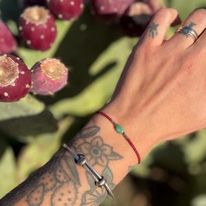 May include: A silver bracelet with a knot design and a red beaded bracelet with a green stone. The bracelets are worn on a tattooed arm, with a background of cactus fruit and a cactus plant.