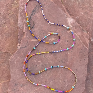 Puede incluir: Un collar de cuentas de colores con pequeñas cuentas en tonos azules, amarillos, naranjas, rosas y turquesas. El collar está sobre una superficie de piedra de color marrón rojizo.