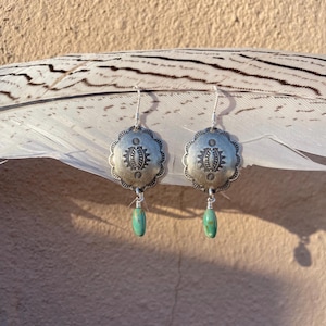 May include: A pair of silver earrings with a scalloped edge and a central embossed design. Each earring has a turquoise-colored teardrop-shaped stone dangling below. The earrings are displayed on a large white feather.