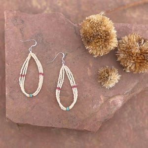 May include: Cream-colored beaded earrings with a teardrop design. The earrings feature silver hooks, with sections of magenta and turquoise beads. The earrings are displayed on a reddish-brown stone surface, with dried, tan-colored flowers.