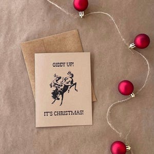Puede incluir: Una tarjeta de Navidad con una ilustración de vaquero y caballo salvaje, el texto dice "Giddy Up! It's Christmas!". La tarjeta está sobre un sobre marrón, con adornos navideños rojos y cuerda.