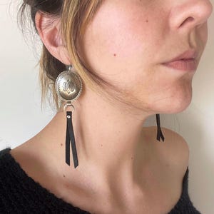 以下が含まれることがあります： 装飾的な花の模様と黒い革のフリンジが付いた、銀色の楕円形のイヤリング。イヤリングは、ヴィンテージルックのウェスタン風の美学を持っています。イヤリングは、ステートメントピースです。