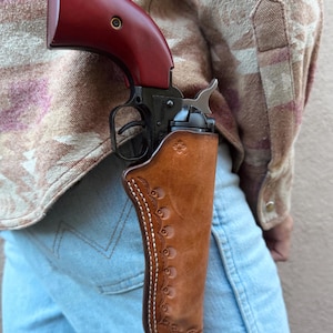 May include: A single-action revolver with a dark metal frame and a smooth, deep red grip, housed in a tan leather holster. The holster features intricate floral-style tooling along the edge, white contrast stitching, and a small stamped sun icon.