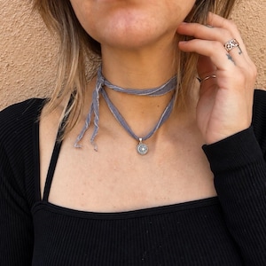 May include: A grey fabric choker necklace featuring a silver-toned circular pendant with a starburst design. The necklace is tied in a knot at the front, with two loose ends hanging down, and is worn around the neck over a black long-sleeve shirt.