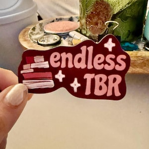 Könnte beinhalten: Ein weinroter Aufkleber mit den rosa Worten "endless + TBR", rosa Sternakzenten und einem Bücherstapel. Der Aufkleber wird von einer Hand gehalten.