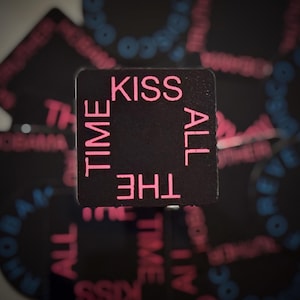May include: A black square with the words "KISS ALL THE TIME" in pink lettering. The phrase is arranged around the edges of the square. Other similar squares are in the background.