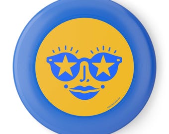 Può includere: Un frisbee blu e giallo con un design a forma di viso sorridente con occhiali da sole a forma di stella. Il viso è giallo con dettagli blu, tra cui gli occhiali da sole, le labbra e le ciglia. Il frisbee ha un bordo blu e il testo "GIRLS STREET BEACH CLUB".