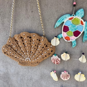 Könnte beinhalten: Eine braune, gehäkelte Handtasche in Muschelform mit goldener Kette und Perlenakzenten. Im Hintergrund hängt ein türkisfarbener Schildkröten-Windspiel mit bunten Mosaikdetails und Muscheldekorationen.