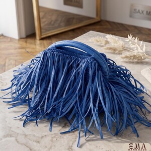 May include: A vibrant blue handbag featuring a dense layer of long, thin fringe strips covering the exterior. The bag has a rounded, structured top handle and a crocheted base construction. The bottom right corner displays the text EMA HANDMADE.