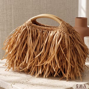 May include: A tan handbag featuring a dense layer of long, thin fringe strips covering the exterior. The bag has a rounded shape with two smooth, light-coloured wooden handles attached to the top. The text EMA HANDMADE is visible in the corner.