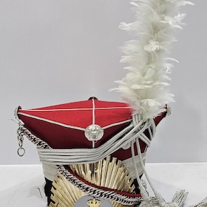 Puede incluir: Un sombrero militar rojo y negro con ribete plateado, un emblema dorado y una gran pluma blanca. El sombrero presenta una cadena y borla plateadas decorativas. El emblema tiene una corona y la letra "N".
