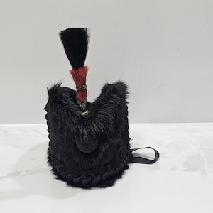 May include: A black fur bag with a black tassel and red accents. The bag has a small handle and a round, black base. The tassel is attached to the top of the bag with a metal ring.