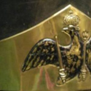 May include: Close-up of a black military hat with a gold-colored emblem. The emblem features a crowned eagle holding a scepter. The hat has a black fur trim. The hat is likely from a historical period.