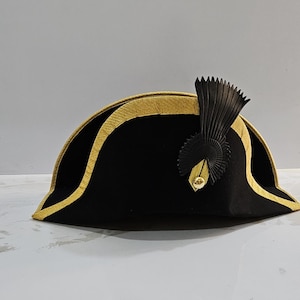 Puede incluir: Un sombrero bicornio negro con ribete dorado y un penacho decorativo negro. El sombrero tiene un ala curva y una corona redondeada. El ribete dorado sigue el borde del ala y el contorno de la corona.