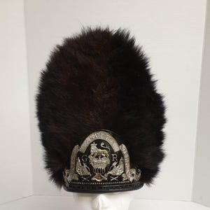 May include: A tall, dark brown fur hat with a decorative silver emblem. The emblem features a crown, lion, and the letters "G" and "R". The hat is likely a ceremonial or military style.