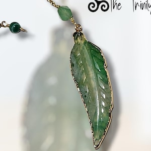 May include: A gold-toned necklace featuring a green leaf pendant with gold trim. The leaf has detailed veining and is suspended from a delicate chain. Additional green beads accent the chain. The text "The Trinity" is in the upper right corner.