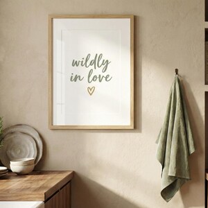 Könnte beinhalten: Gerahmter Druck mit den Worten "wildly in love" in einer Schreibschrift, mit einem goldenen Herz. Der Druck hängt an einer beigefarbenen Wand über einer Holz-Arbeitsplatte. Ein grünes Handtuch hängt an einem Haken. Ein Regal enthält Bücher und einen Kupferkessel.