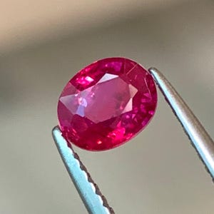 Può includere: Una vibrante gemma di rubino taglio ovale, tenuta da una pinzetta d'argento. Il rubino mostra una tonalità rosso intenso con sfaccettature interne che riflettono la luce. La pinzetta è a fuoco, evidenziando la chiarezza.