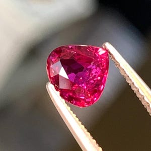 Puede incluir: Una gema roja vibrante, facetada y en forma de corazón, es sostenida por unas pinzas plateadas. El color de la gema varía del magenta oscuro al rosa brillante, con múltiples facetas que capturan la luz. El fondo está borroso, enfatizando el brillo de la gema.