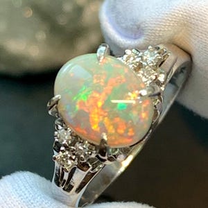 Könnte beinhalten: Silberring mit einem ovalen Opal, der farbige Lichtreflexe zeigt. Der Opal ist von kleineren, klaren Edelsteinen umgeben. Der Ring wird von einer behandschuhten Hand gehalten, was den Schmuck hervorhebt.