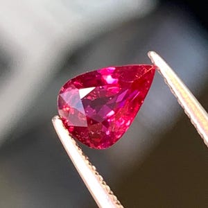 May include: A faceted, pear-shaped, deep red gemstone is held by tweezers. The gem has a brilliant cut, reflecting light. The tweezers are silver-coloured, and the background is blurred.