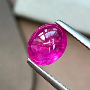 Op de afbeelding: Een levendige, ovaalvormige edelsteen in cabochon-slijpvorm met een diepe magenta-roze tint. De steen wordt stevig vastgehouden door een zilverkleurige metalen pincet, wat het gladde, gepolijste en reflecterende oppervlak onder fel licht toont.