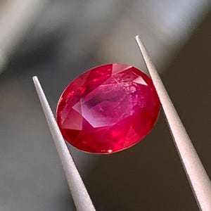 Op de afbeelding: Een levendige, ovaal geslepen robijn met een dieprode kleur en een veelzijdig oppervlak, vastgehouden door een metalen pincet. De edelsteen vertoont interne helderheid en lichtreflectie over het gepolijste, symmetrische en gefacetteerde bovenoppervlak.