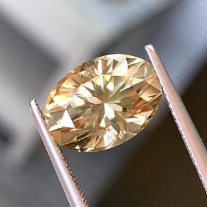 May include: A faceted, oval-cut gemstone held by tweezers. The gem has a warm, golden-yellow hue and a brilliant, multi-faceted surface. The tweezers are silver-colored and hold the gem securely, showcasing its clarity and cut.