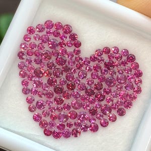 Pode incluir: Um conjunto de pequenas gemas rosas e redondas dispostas em forma de coração. As pedras preciosas são apresentadas num recipiente quadrado branco com um interior branco. As gemas são cortadas para refletir a luz.