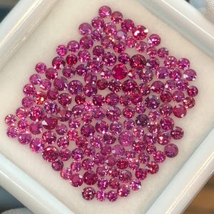 Puede incluir: Una colección de pequeñas gemas rosas, redondas y facetadas, exhibidas en un contenedor cuadrado blanco. Las gemas varían en tamaño y tono, desde rosa claro hasta magenta intenso. El contenedor se encuentra sobre un fondo blanco suave.