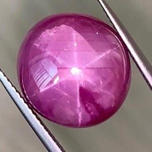 以下が含まれることがあります： 銀色のピンセットで保持された、研磨された丸いピンク色の宝石。6条の星の効果があります。宝石は光沢のある表面を持ち、光を反射してスターバーストパターンを作り出しています。ピンセットは斜めに配置され、宝石をフレーミングしています。