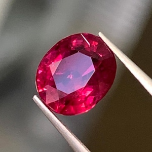 Puede incluir: Una gema de rubí de corte ovalado, con una saturación de color rojo intenso y superficies facetadas que reflejan la luz. La gema está sujeta por unas pinzas de metal, mostrando su claridad transparente y sus bordes pulidos.
