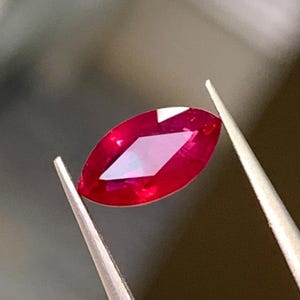 以下が含まれることがあります： 鮮やかな深紅の色合いとファセットカットが施されたマーキスカットの宝石。銀色の金属製ピンセットで保持されており、細長く尖った形状と透明な素材を通して見える内部の光の反射が特徴的です。